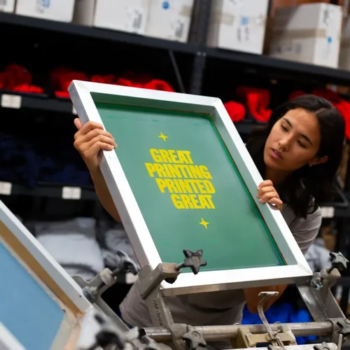 A person inspects a screen printing frame with the words "GREAT PRINTING PRINTED GREAT" in a workshop setting.