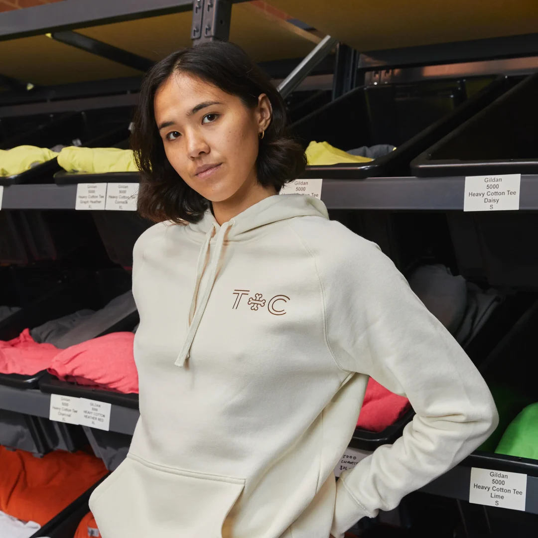 A person is standing in front of a range of t-shirts wearing a grey hoodie with a custom logo on it