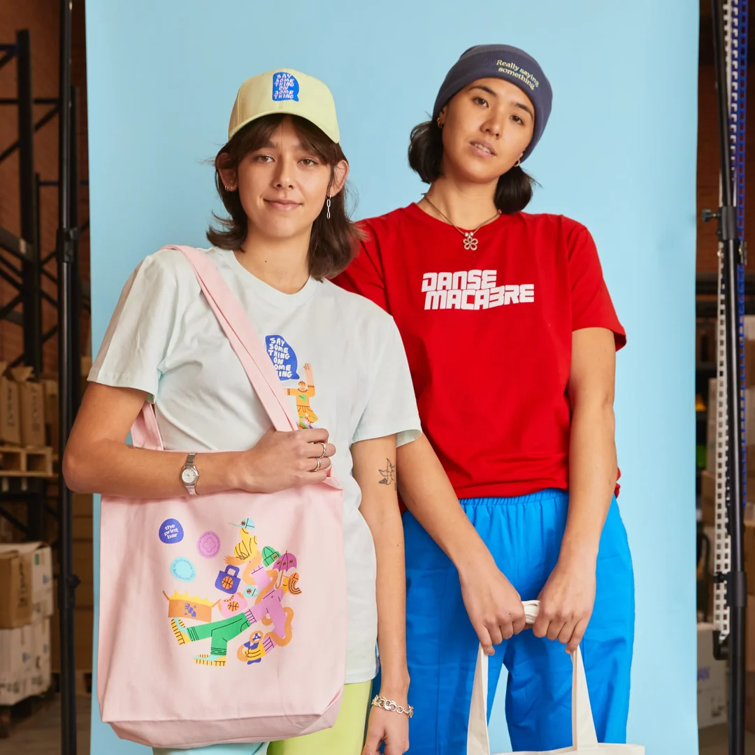Two people in custom t-shirts, embroidered hats and beanies with personalised tote bags stand smiling