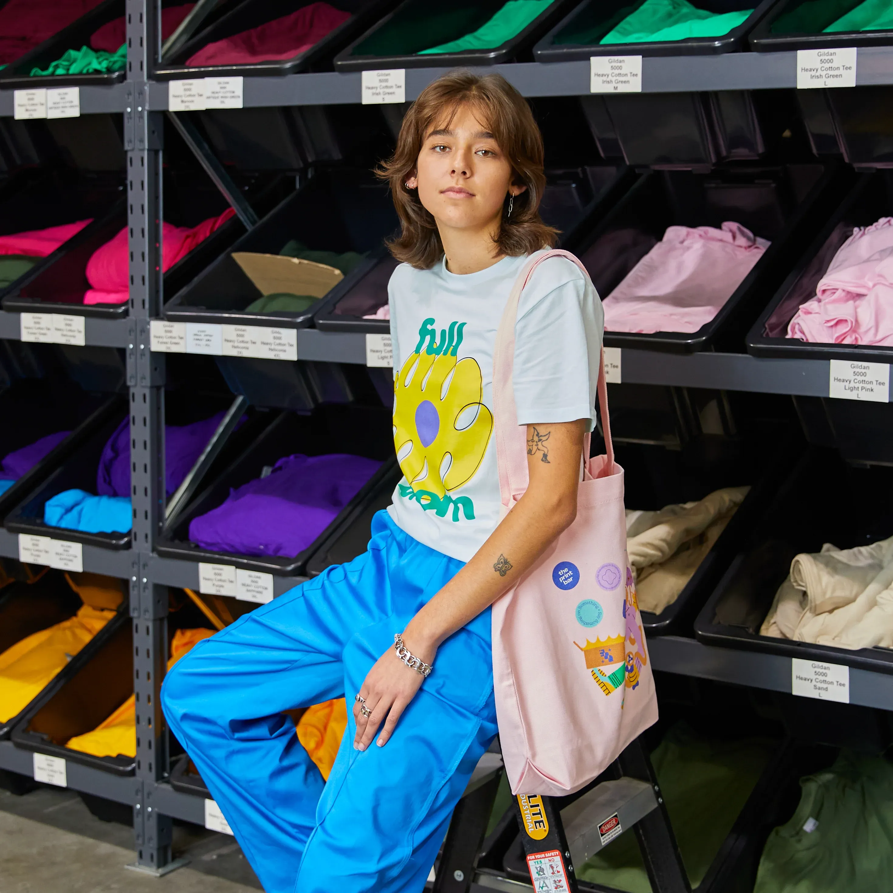 Person sits on a chair in front of a t-shirt storage wearing a t-shirt and tote bag with custom logos