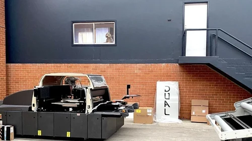 In a warehouse setting a large industrial printer sits, in the backgrounds sits a window with a small dog looking through it