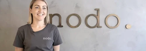 A woman standing and smiling wears a shirt that reads 'nodo' stands in front of a concrete wall that reads 'nodo.'