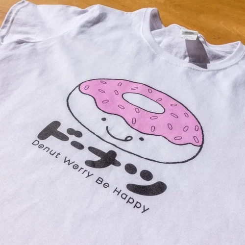 A T-shirt lays on a table that reads 'Donut Worry Be Happy' with a donut cartoon